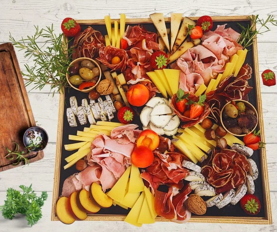Planche en bois garnie de fromages, saucisson, jambon sec, olives, cornichons et pain, avec deux verres de vin
